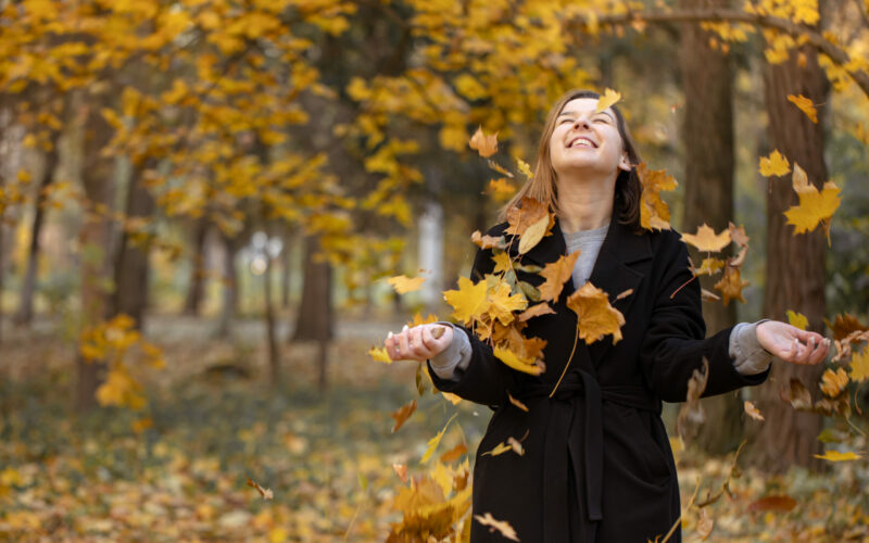 Happy young woman in the forest among autumn leaves on a blurred background. Happy young woman in a black coat in the forest among flying autumn leaves on a blurred background, copy space.